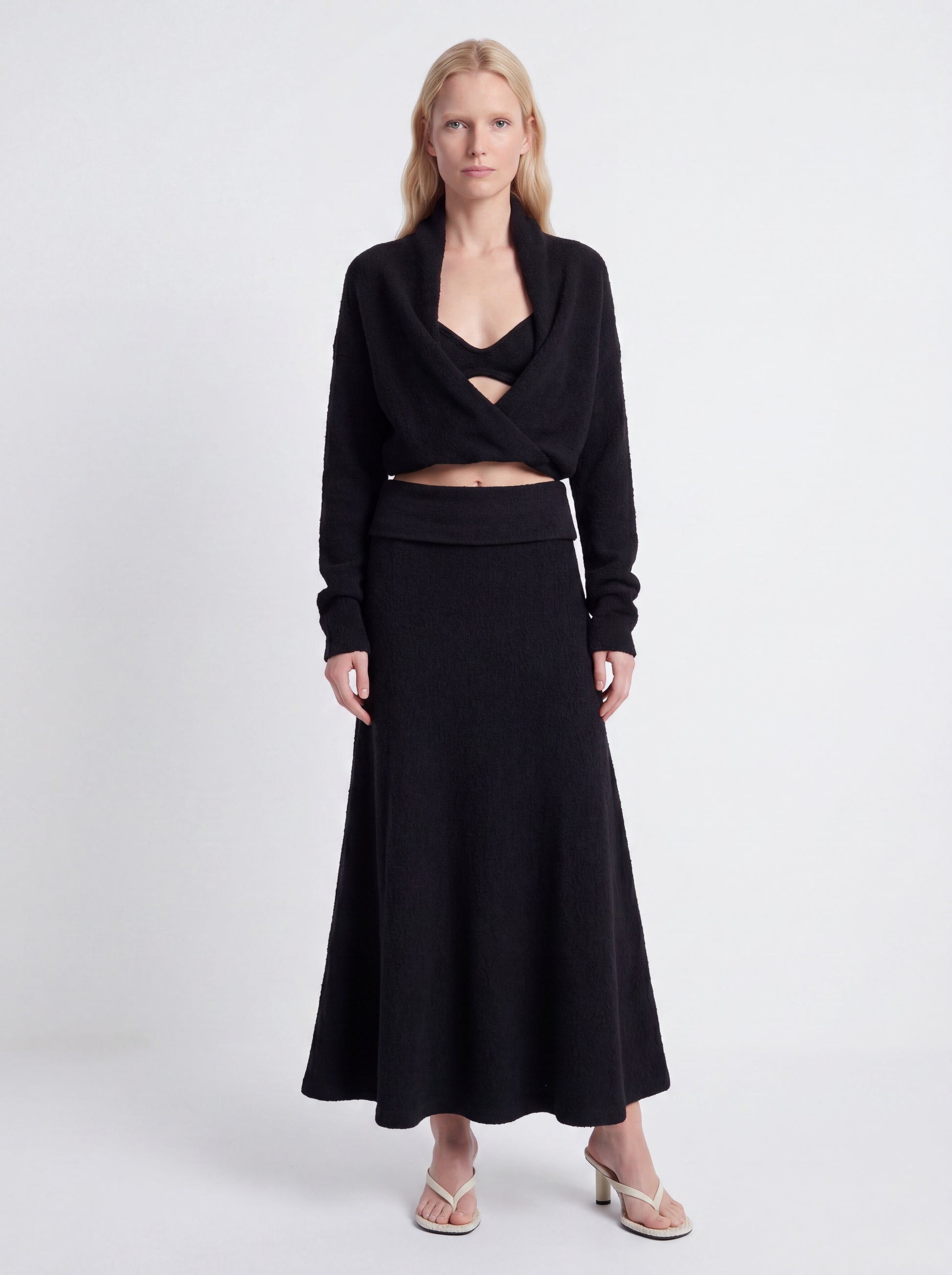 A model wearing a black two-piece knit set consisting of a long-sleeve wrap-style cropped top with a crossover neckline and a matching high-waisted maxi skirt, paired with beige heeled sandals against a light gray studio background.