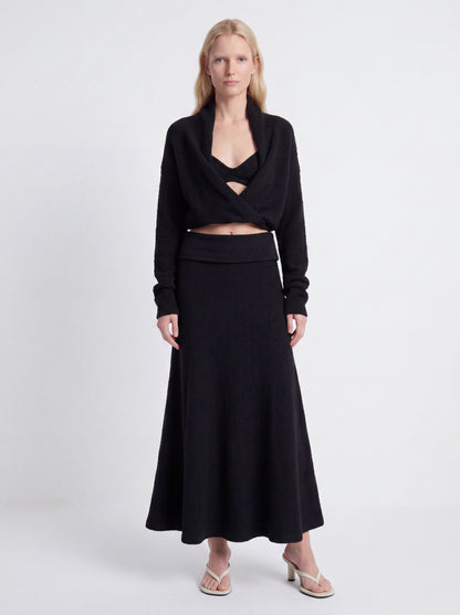 A model wearing a black two-piece knit set consisting of a long-sleeve wrap-style cropped top with a crossover neckline and a matching high-waisted maxi skirt, paired with beige heeled sandals against a light gray studio background.