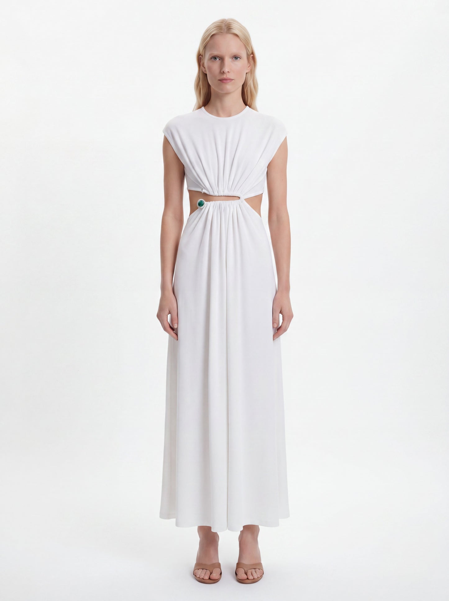 A blonde model wearing a white sleeveless maxi dress with a gathered, pleated bodice, a horizontal cutout at the waist, and a flowing skirt, standing against a plain white studio background.