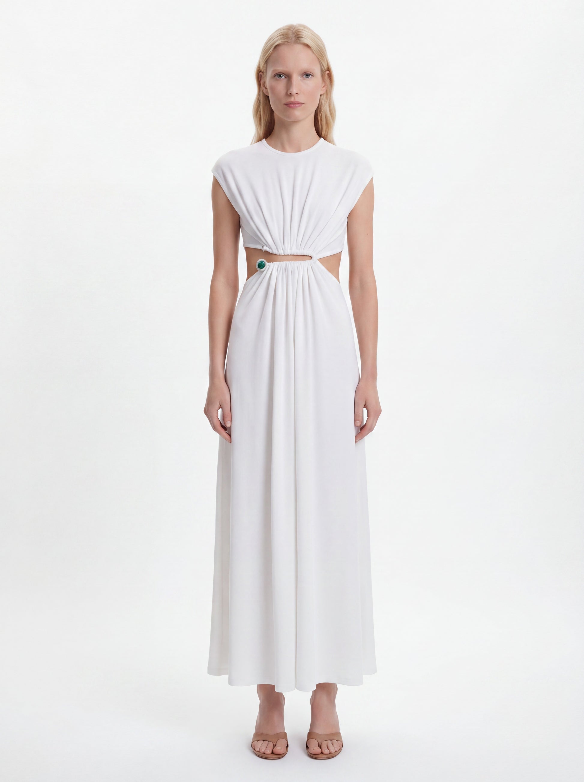 A blonde model wearing a white sleeveless maxi dress with a gathered, pleated bodice, a horizontal cutout at the waist, and a flowing skirt, standing against a plain white studio background.