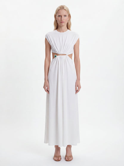 A blonde model wearing a white sleeveless maxi dress with a gathered, pleated bodice, a horizontal cutout at the waist, and a flowing skirt, standing against a plain white studio background.