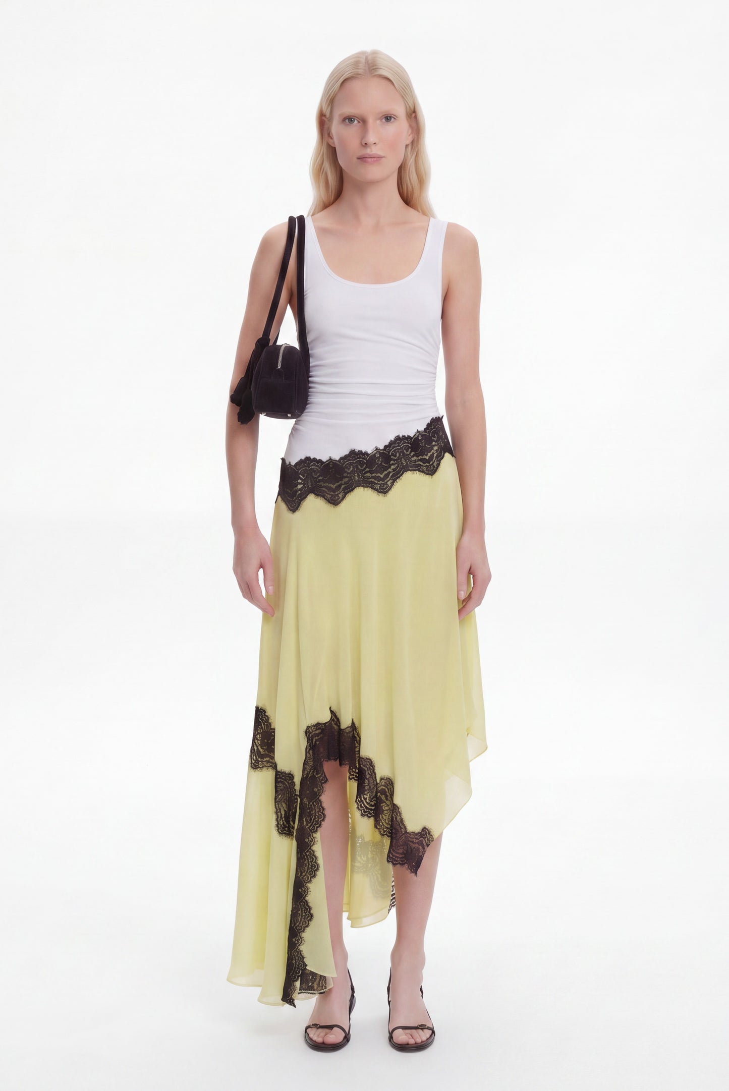 A model stands against a white studio background wearing a white ribbed tank top, a flowing pale yellow skirt with black lace trim, and black sandals, while carrying a black shoulder bag.