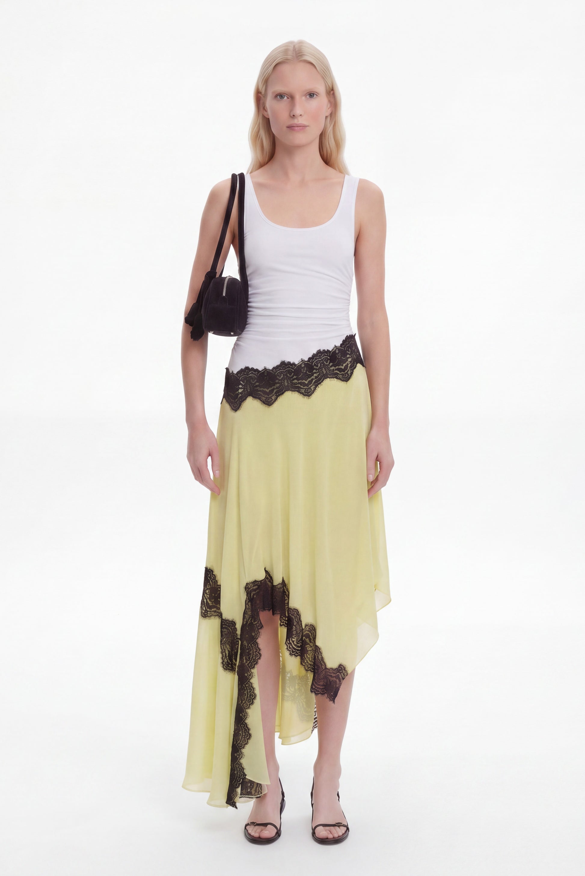 A model stands against a white studio background wearing a white ribbed tank top, a flowing pale yellow skirt with black lace trim, and black sandals, while carrying a black shoulder bag.