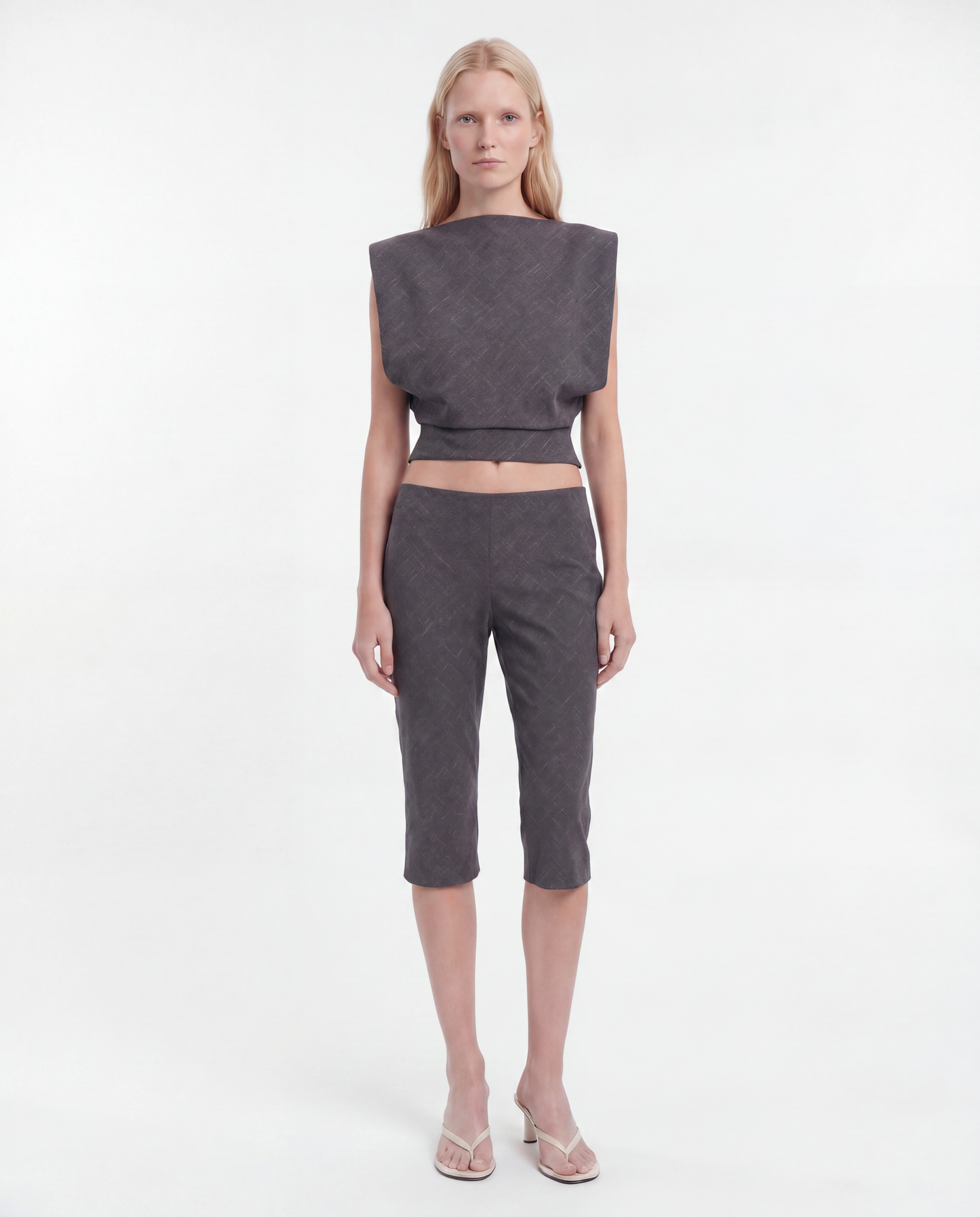 A model stands against a plain white studio background wearing a dark gray textured two-piece set consisting of a sleeveless cropped top with wide shoulders and matching cropped pants, paired with clear high-heeled sandals.