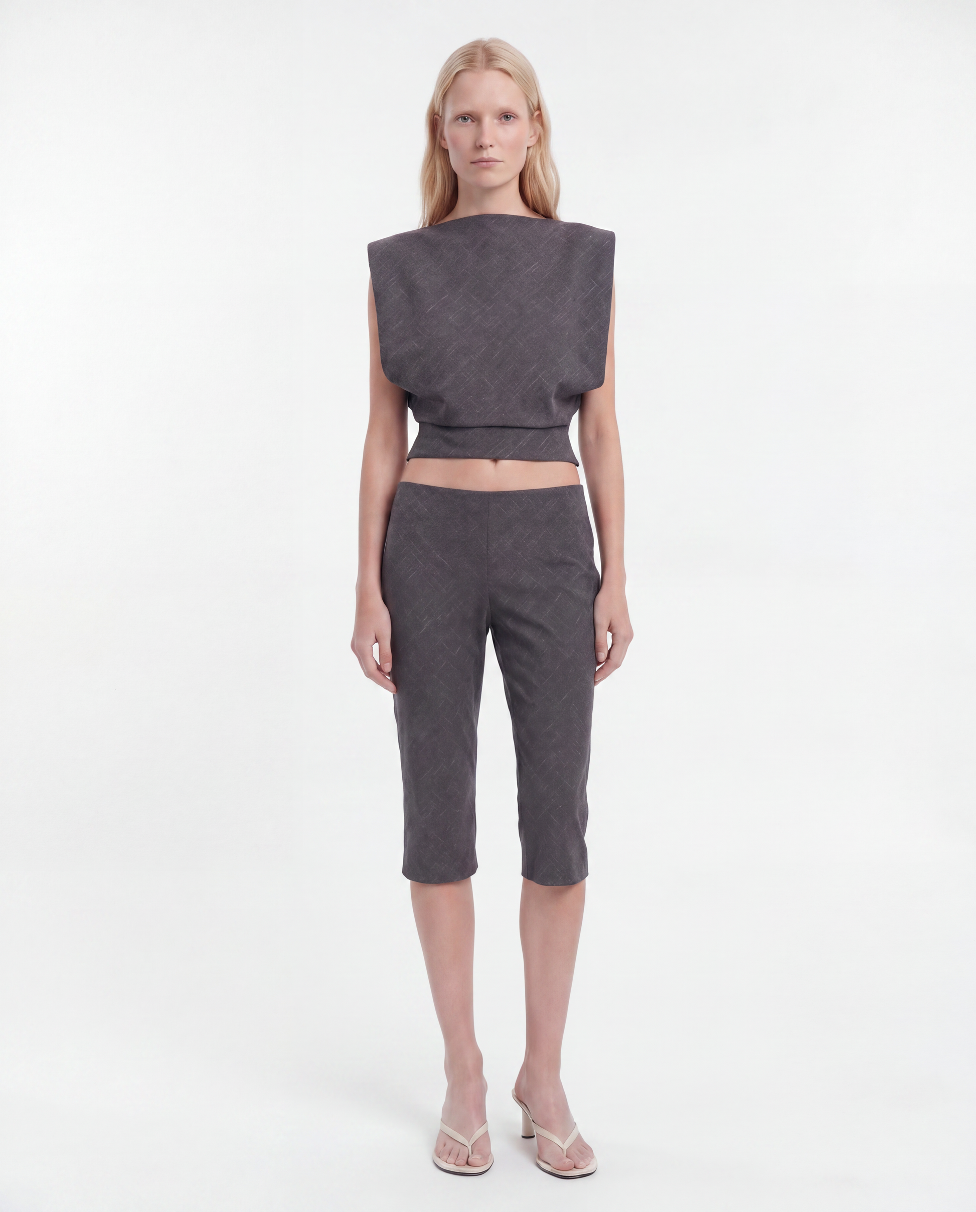 A model stands against a plain white studio background wearing a dark gray textured two-piece set consisting of a sleeveless cropped top with wide shoulders and matching cropped pants, paired with clear high-heeled sandals.