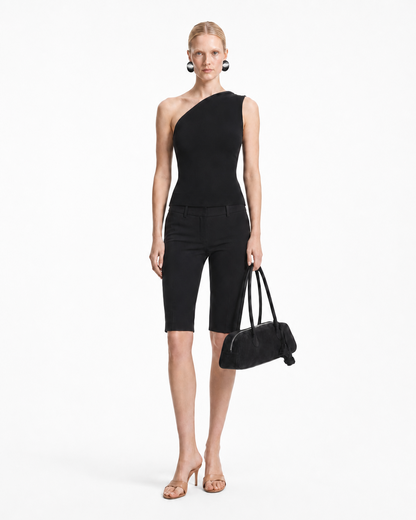 A model stands against a plain white studio background wearing a black one-shoulder knit jumpsuit with black cropped shorts, nude high-heeled sandals, and holding a black handbag.